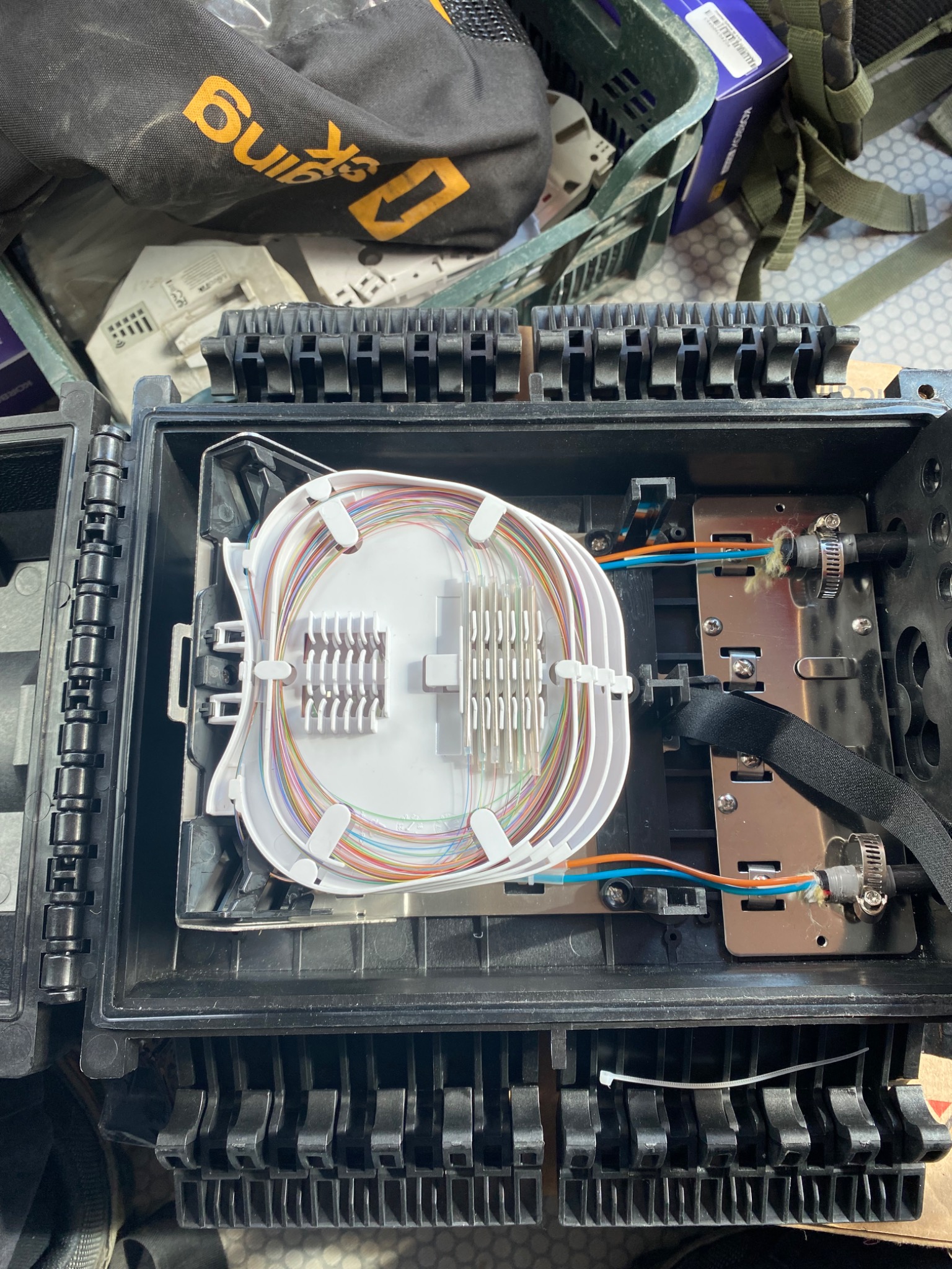 Otwarta czarna skrzynka telekomunikacyjna z widocznym światłowodem ułożonym wewnątrz, przewody w kolorach pomarańczowym i niebieskim podłączone do metalowej płyty, widoczny pasek mocujący i biały...