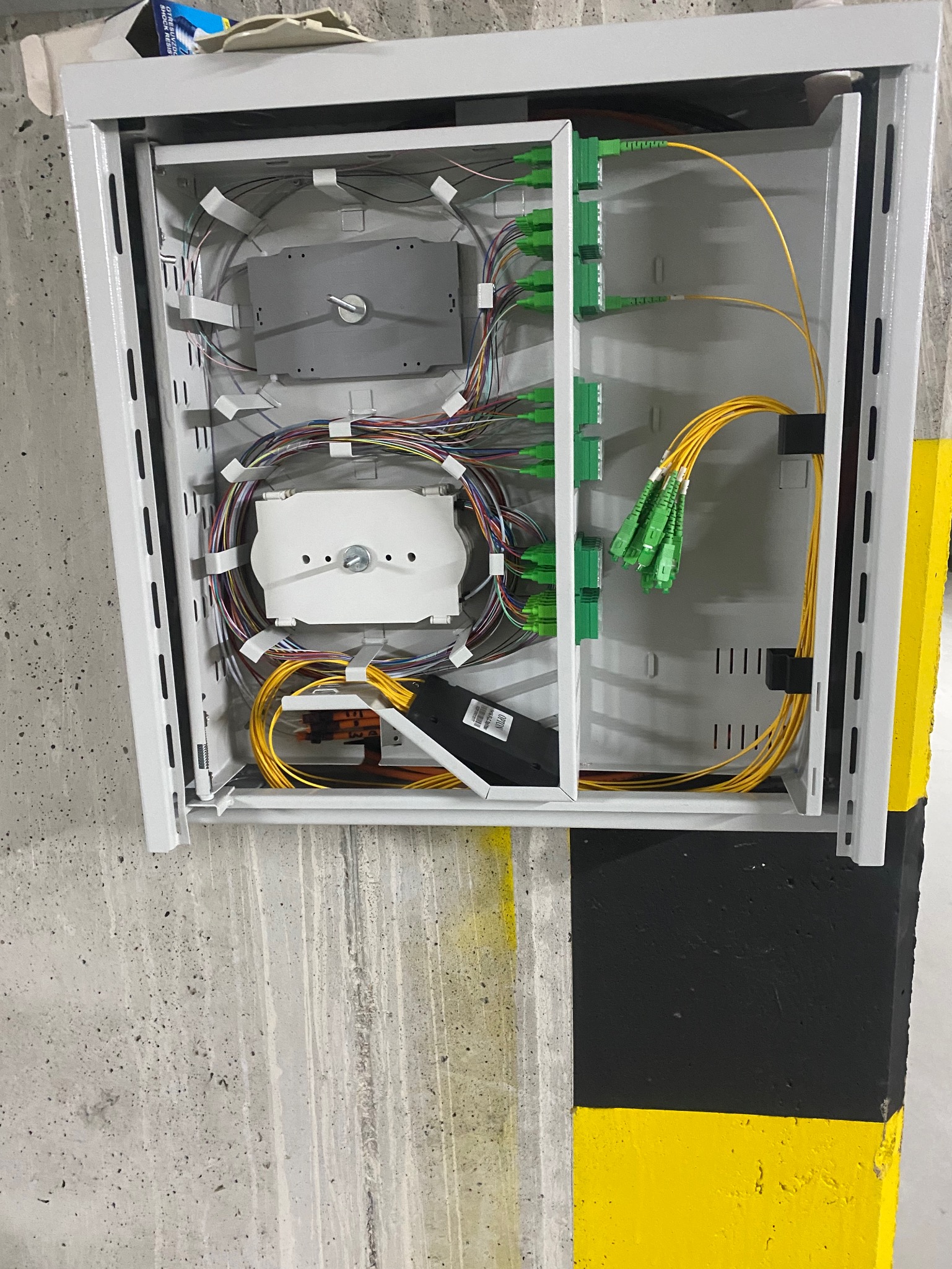 Otwarta, metalowa szafka telekomunikacyjna z rozdzielnicą światłowodową, kable światłowodowe w kolorze żółtym i zielone złącza, zamontowana na betonowej ścianie z czarno-żółtym oznaczeniem.