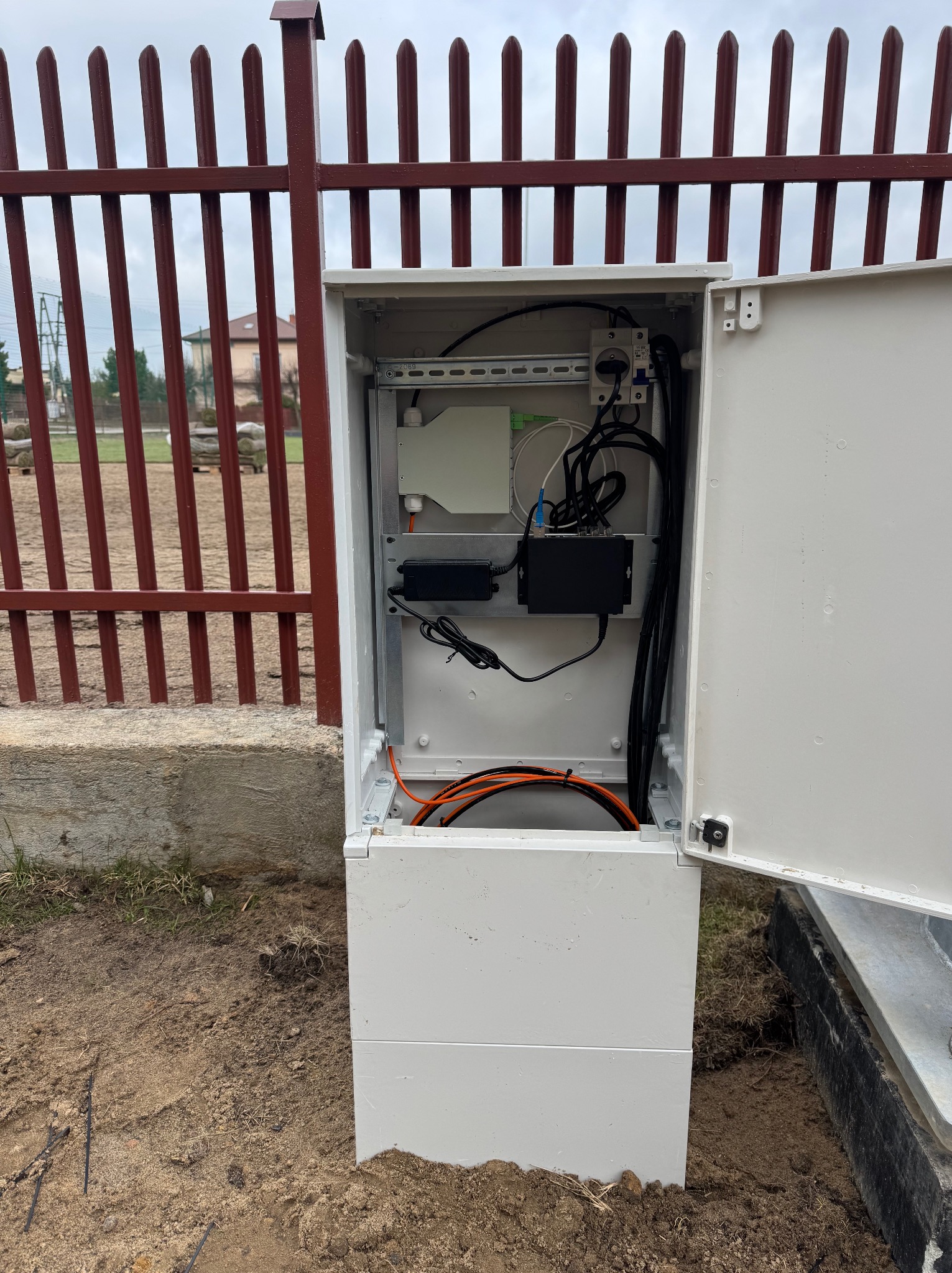 Otwarta biała szafka telekomunikacyjna z okablowaniem i urządzeniami, umieszczona na zewnątrz przy metalowym ogrodzeniu.