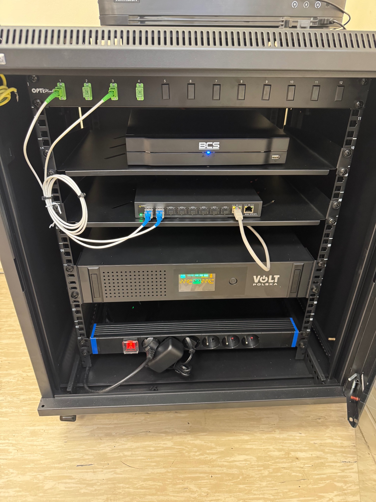 Szafa rack z zainstalowanymi urządzeniami sieciowymi: patch panel, switch, zasilacz awaryjny VOLT Polska i listwa zasilająca. Widoczne kable światłowodowe i ethernet.