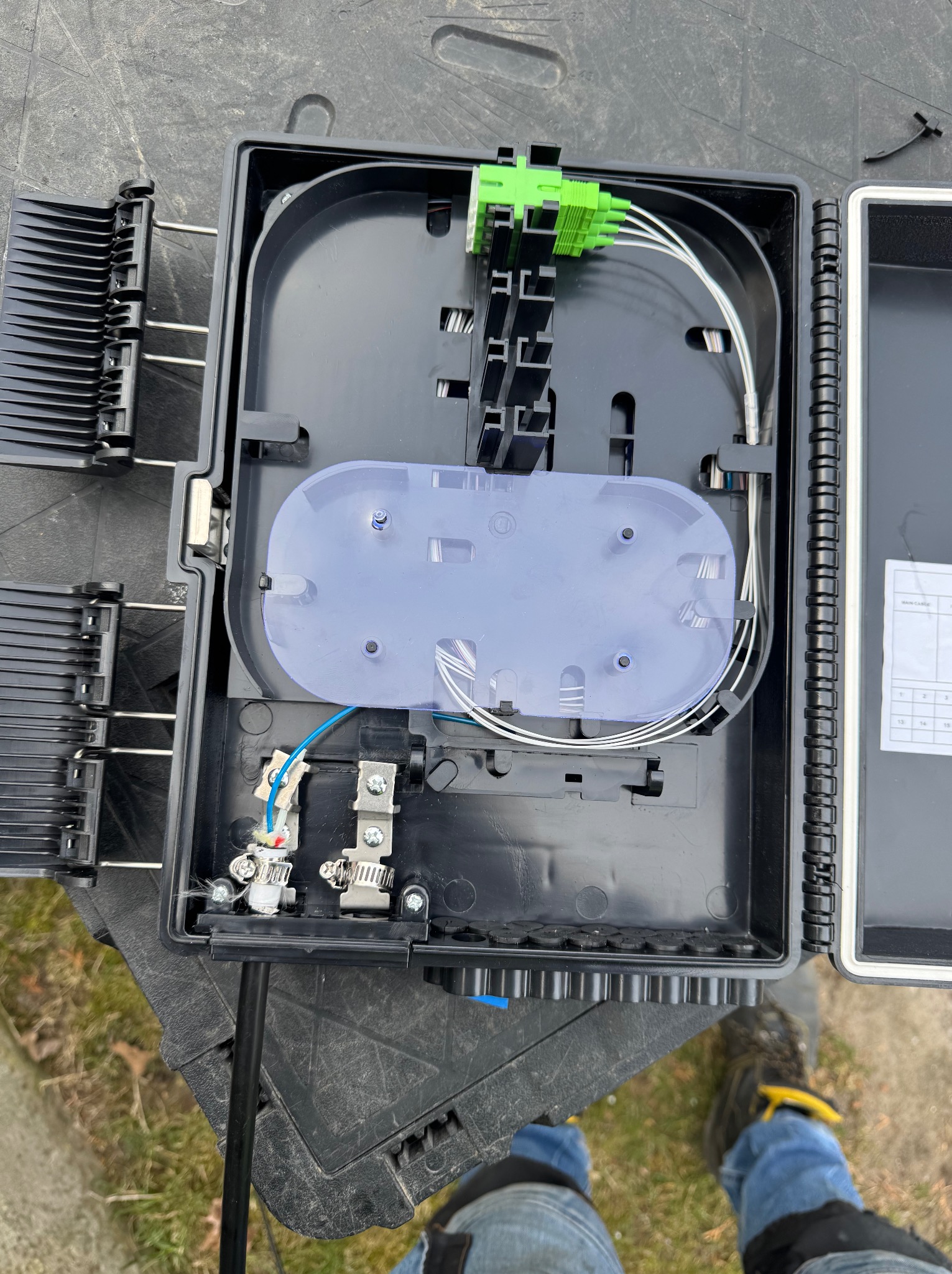 Otwarta, czarna skrzynka telekomunikacyjna z widocznymi światłowodami, złączkami i elementami montażowymi. Widoczne kable i złącza wewnątrz. Widok z góry.