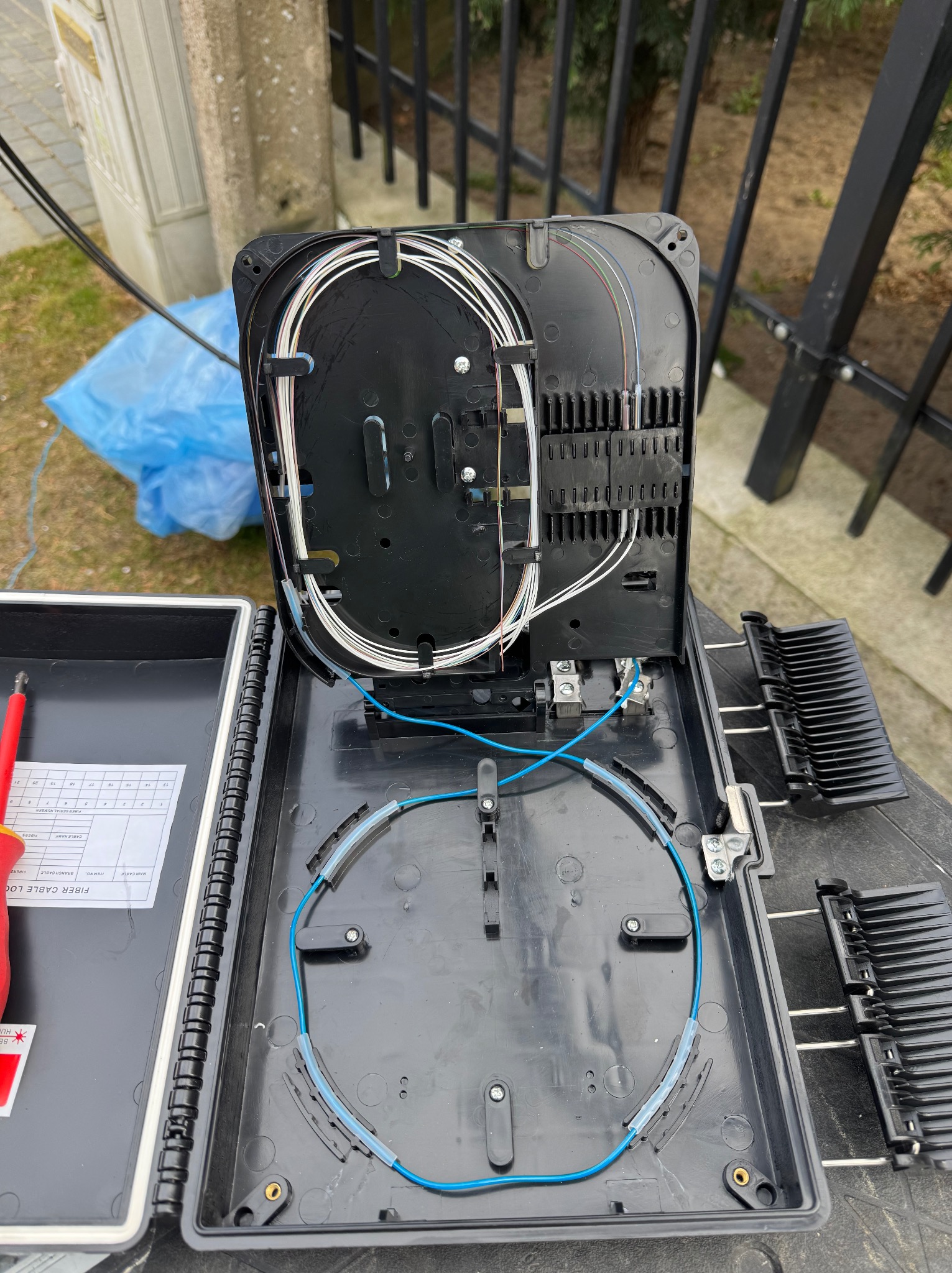 Otwarta skrzynka telekomunikacyjna z widocznymi kablami światłowodowymi i narzędziami. Układanie kabli w czarnej skrzynce na zewnątrz budynku.