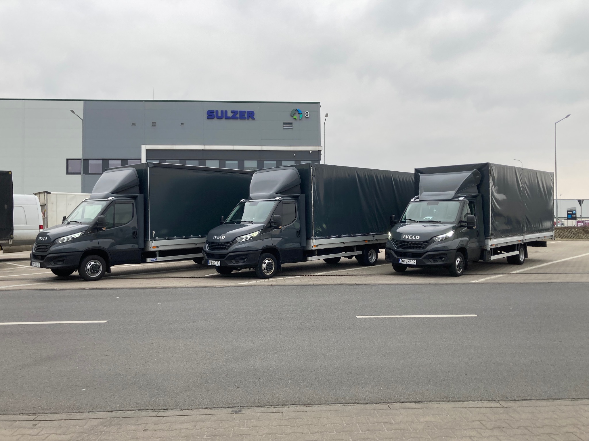 Trzy identyczne, grafitowe busy dostawcze marki Iveco z plandekami zaparkowane równolegle przed budynkiem firmy Sulzer w pochmurny dzień.