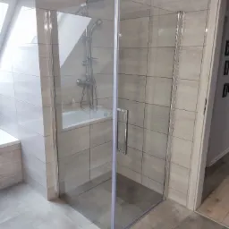 Szklana kabina prysznicowa z chromowanymi profilami, zamontowana w narożniku łazienki z szarymi płytkami, widoczna bateria prysznicowa i wanna w tle.