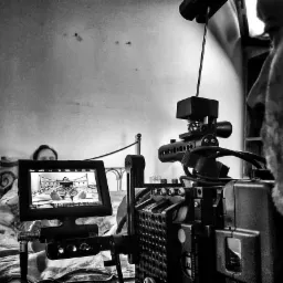 Czarno-białe ujęcie zza kamery filmowej, na ekranie podgląd osoby leżącej w łóżku i czytającej książkę, w tle fragment twarzy operatora.