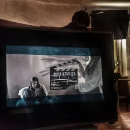 Ekran monitora z czarno-białym nagraniem kobiety i klapsa filmowego, w tle oświetlenie studyjne i sylwetki osób.