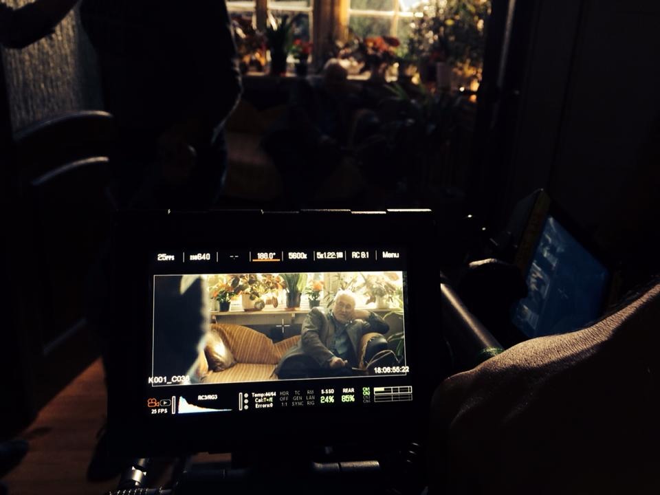 Ekran monitora filmowego z podglądem sceny: starszy mężczyzna siedzący na kanapie w salonie z roślinami w tle, ustawienia kamery widoczne na wyświetlaczu.