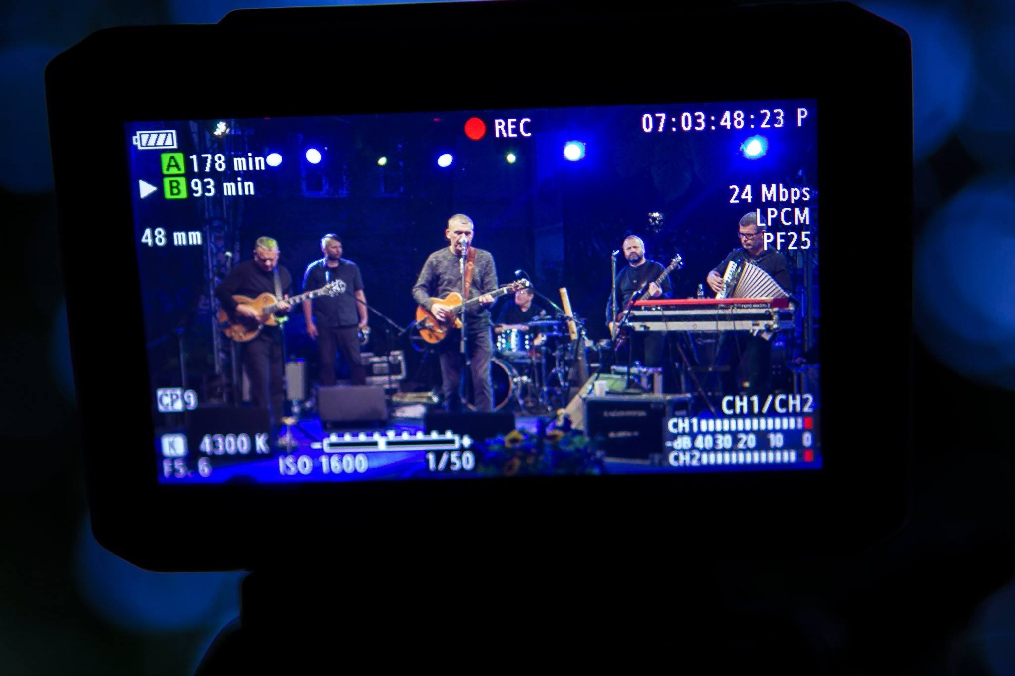 Ekran kamery rejestrującej występ zespołu muzycznego na scenie, widoczne ustawienia nagrywania i wskaźniki audio.