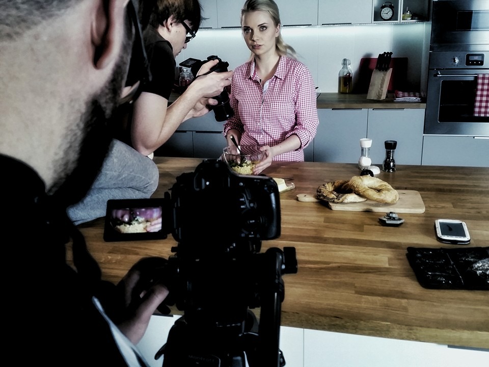 Ekipa filmowa nagrywa kobietę przygotowującą jedzenie w kuchni; widoczny profesjonalny sprzęt kamerowy i oświetlenie.