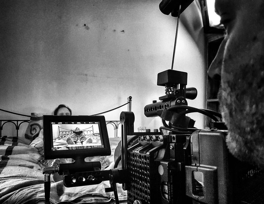 Czarno-białe ujęcie zza kamery filmowej, na ekranie podgląd osoby leżącej w łóżku i czytającej książkę, w tle fragment twarzy operatora.