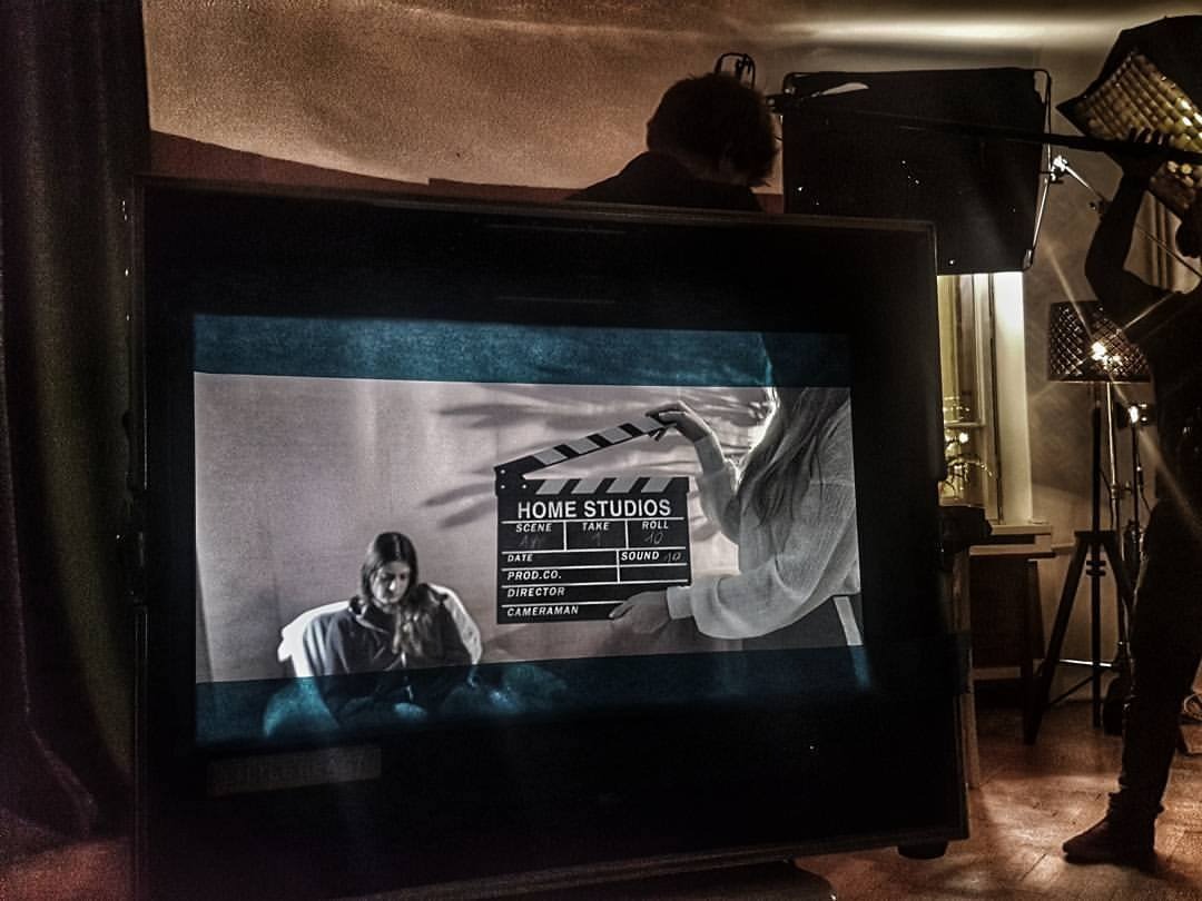 Ekran monitora z czarno-białym nagraniem kobiety i klapsa filmowego, w tle oświetlenie studyjne i sylwetki osób.