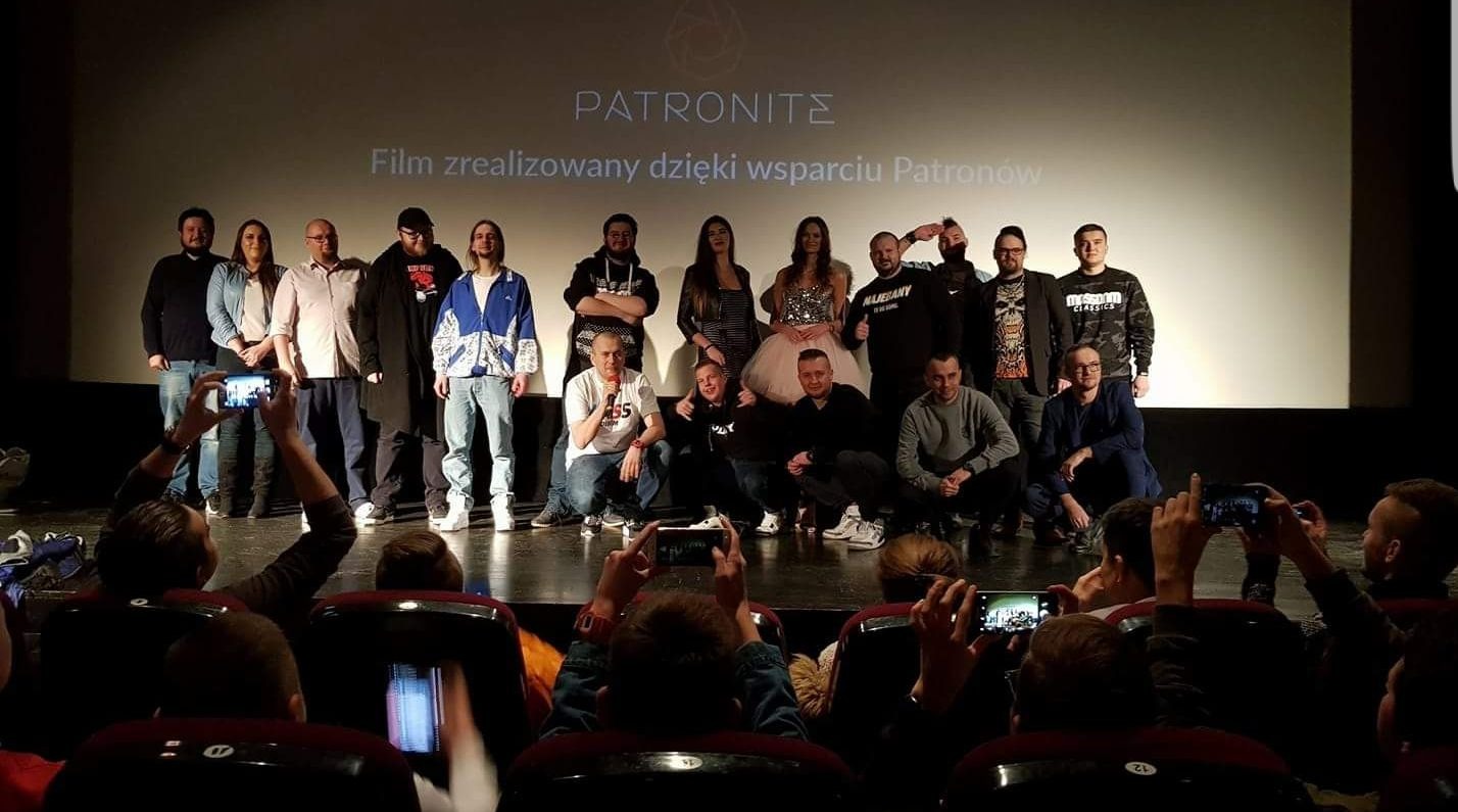 Grupa osób na scenie kina podczas premiery filmu, widzowie filmują wydarzenie telefonami, na ekranie napis 'Film zrealizowany dzięki wsparciu Patronów'.