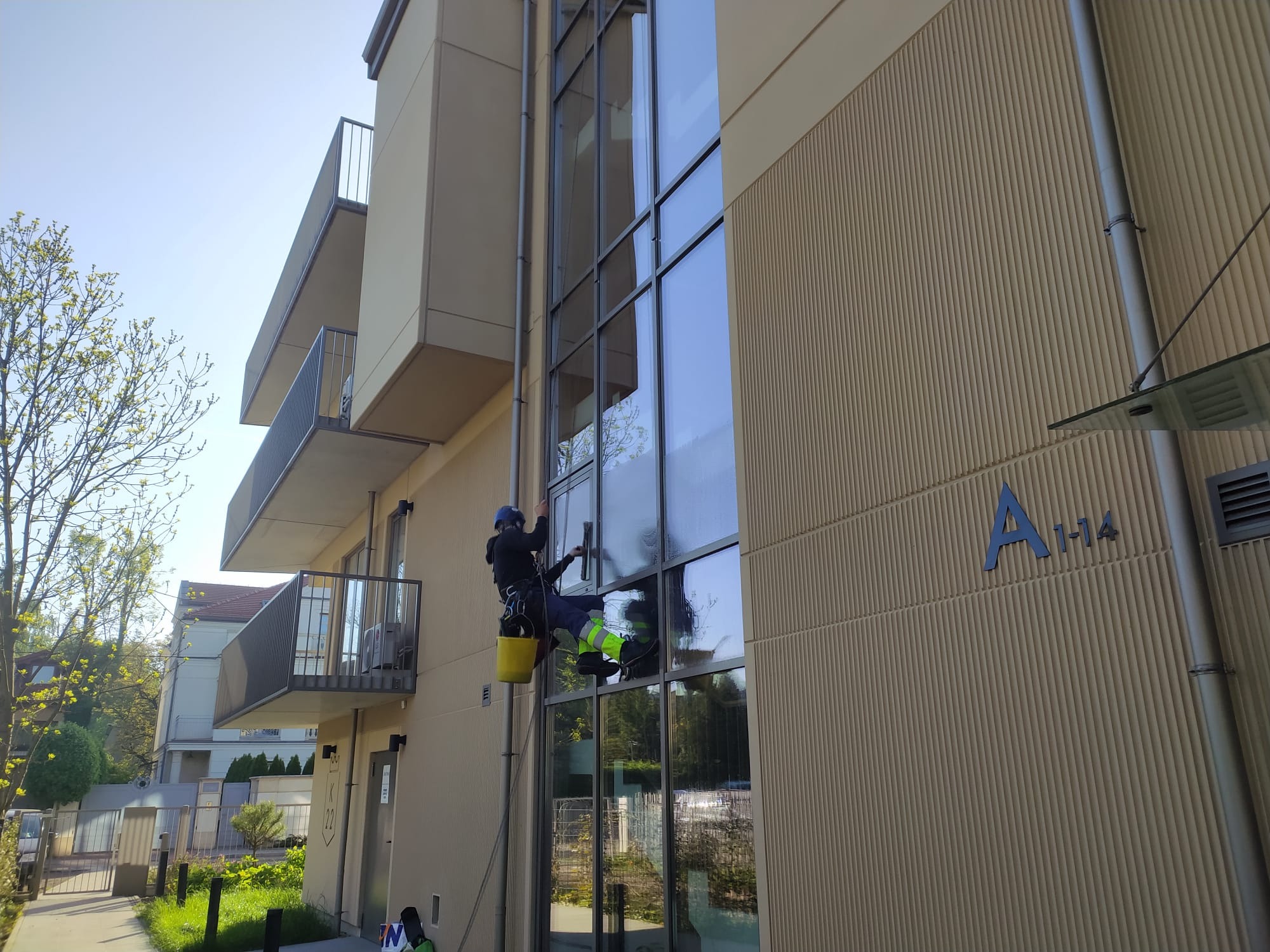 Pracownik na wysokości myje okna nowoczesnego budynku z żółtym wiadrem. Widoczne odbicia w szybach i fragment budynku z balkonami. Słoneczny dzień.