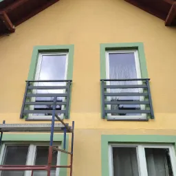 Żółta elewacja domu z dwoma oknami z szarą balustradą i zieloną ramą, widok z dołu, fragment rusztowania. Balustrady okienne zabezpieczające przed wypadnięciem.