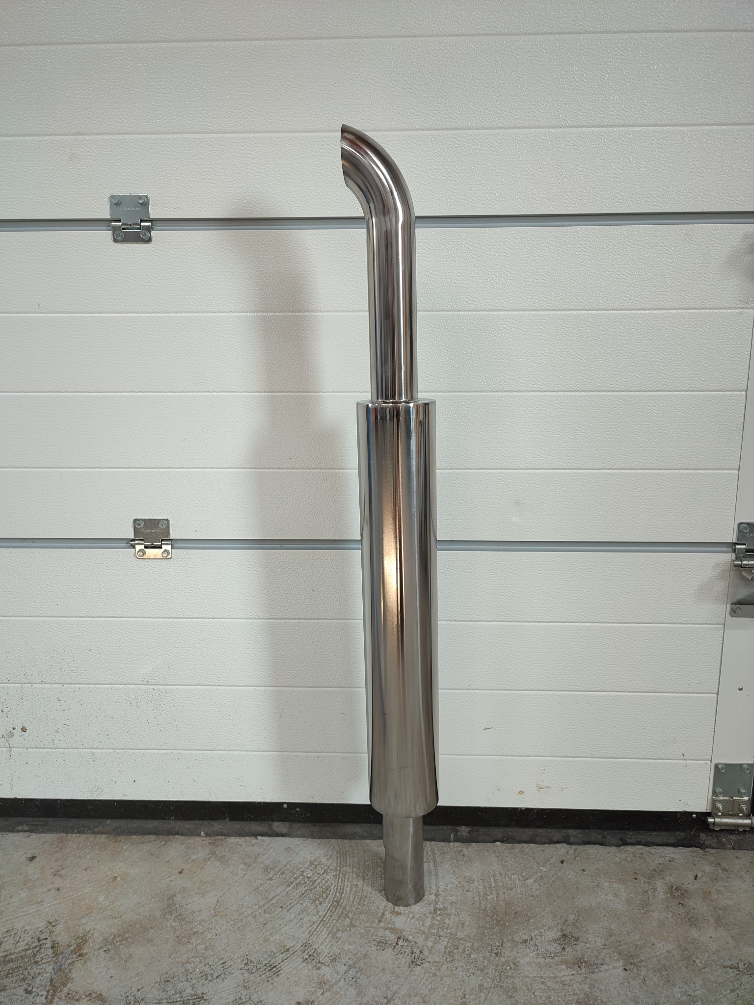Błyszcząca, metalowa rura wygięta w łuk, ustawiona pionowo na tle białej bramy garażowej. Widoczne mocowania bramy i betonowa posadzka.