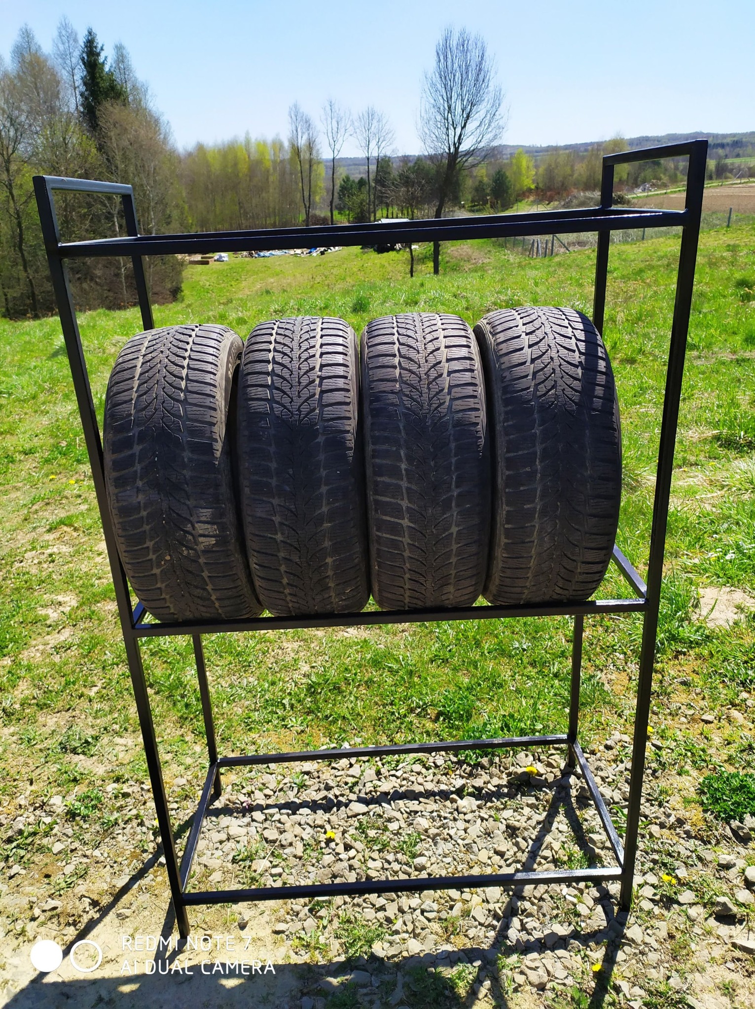 Metalowy stojak na opony, cztery opony ułożone poziomo, tło: trawa i drzewa, widok z zewnątrz, spawana konstrukcja z profili.