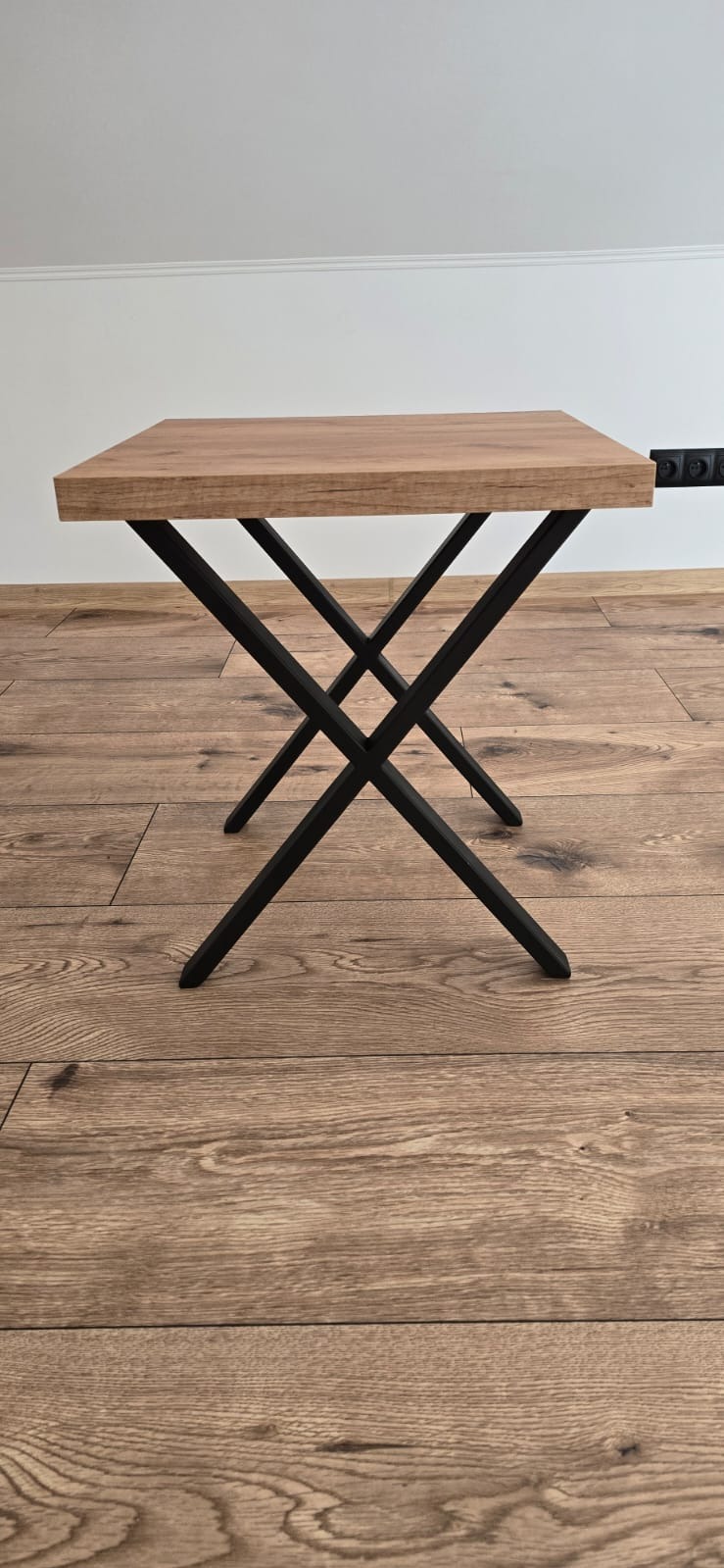 Stolik kawowy z drewnianym blatem i czarnymi, metalowymi nogami w kształcie litery X. Minimalistyczny design, parkiet na podłodze, biała ściana w tle.