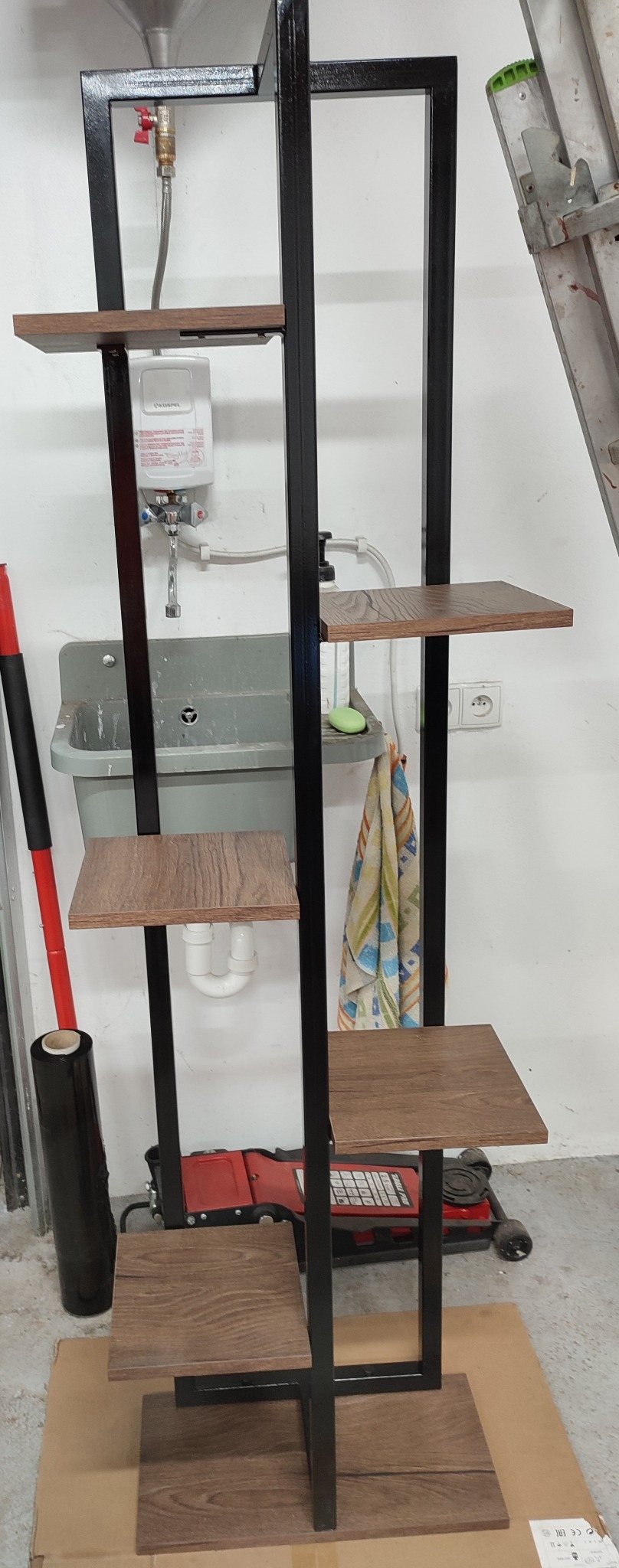 Metalowa konstrukcja z drewnianymi półkami, czarna rama, widoczne spawy, tło warsztatowe z umywalką i narzędziami. Półki z płyty imitującej drewno.