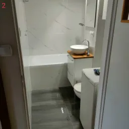 Nowoczesna łazienka z wanną obłożoną białymi płytkami imitującymi marmur, umywalką nablatową na drewnianym blacie, szarą podłogą oraz pralką. Na pralce leży opakowanie proszku do prania.