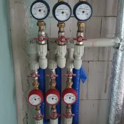 Trzy pary liczników wody (ciepłej i zimnej) zamontowane na rurach w pomieszczeniu o surowych ścianach, z widocznymi zaworami i izolacją termiczną na niektórych rurach.