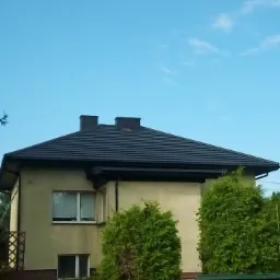 Dom jednorodzinny z nowym, ciemnym dachem z dwoma kominami, widok z poziomu gruntu w słoneczny dzień.