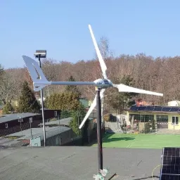 Mała turbina wiatrowa z trzema białymi łopatami, zamontowana na dachu z papy, obok paneli słonecznych. W tle drzewa i boisko sportowe.