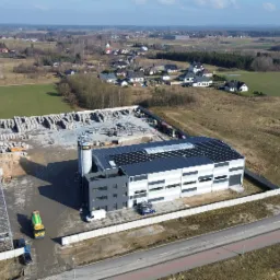 Widok z lotu ptaka na budynek przemysłowy z panelami fotowoltaicznymi na dachu, placem składowym z betonowymi elementami, ciężarówką z betonomieszarką i okolicznym krajobrazem.