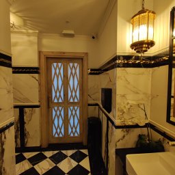 Firma zakładowa - Wnętrze łazienki z marmurowymi płytkami w czarno-białą szachownicę na podłodze, drewnianymi drzwiami z geometrycznym wzorem, umywalką i ozdobną lampą wiszącą.