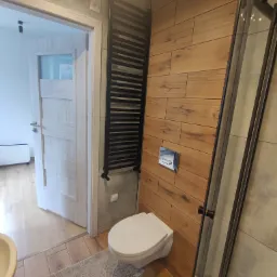 Nowoczesna łazienka z drewnopodobnymi płytkami na ścianie, wiszącą toaletą, czarnym grzejnikiem drabinkowym i kabiną prysznicową z czarnymi ramami.