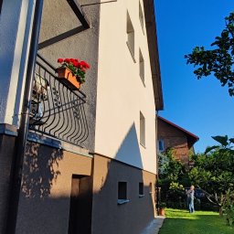 Firma zakładowa - Dwukondygnacyjny dom z jasną elewacją i ciemnobrązowym cokołem, balkon z czerwonymi kwiatami, widok z dołu na tle błękitnego nieba i zielonego trawnika.