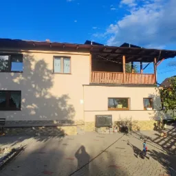 Jasny, ocieplony budynek z drewnianą konstrukcją balkonu na piętrze, widoczny z perspektywy podw&oacute;rza z kostki brukowej, gdzie cień osoby robiącej zdjęcie pada na podłoże.