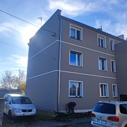 Firma zakładowa - Odnowiony budynek wielorodzinny z szarą elewacją i białymi pasami, widoczne nowe rynny, pracownik z taczką i narzędziami przy budynku, dwa samochody na podwórku, błękitne niebo.