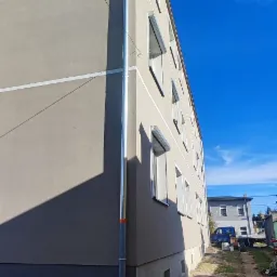 Odnowiona elewacja budynku wielorodzinnego z nową, jasną farbą i orynnowaniem, widoczne okna z roletami zewnętrznymi, dolna część budynku w ciemniejszym odcieniu, błękitne niebo w tle.