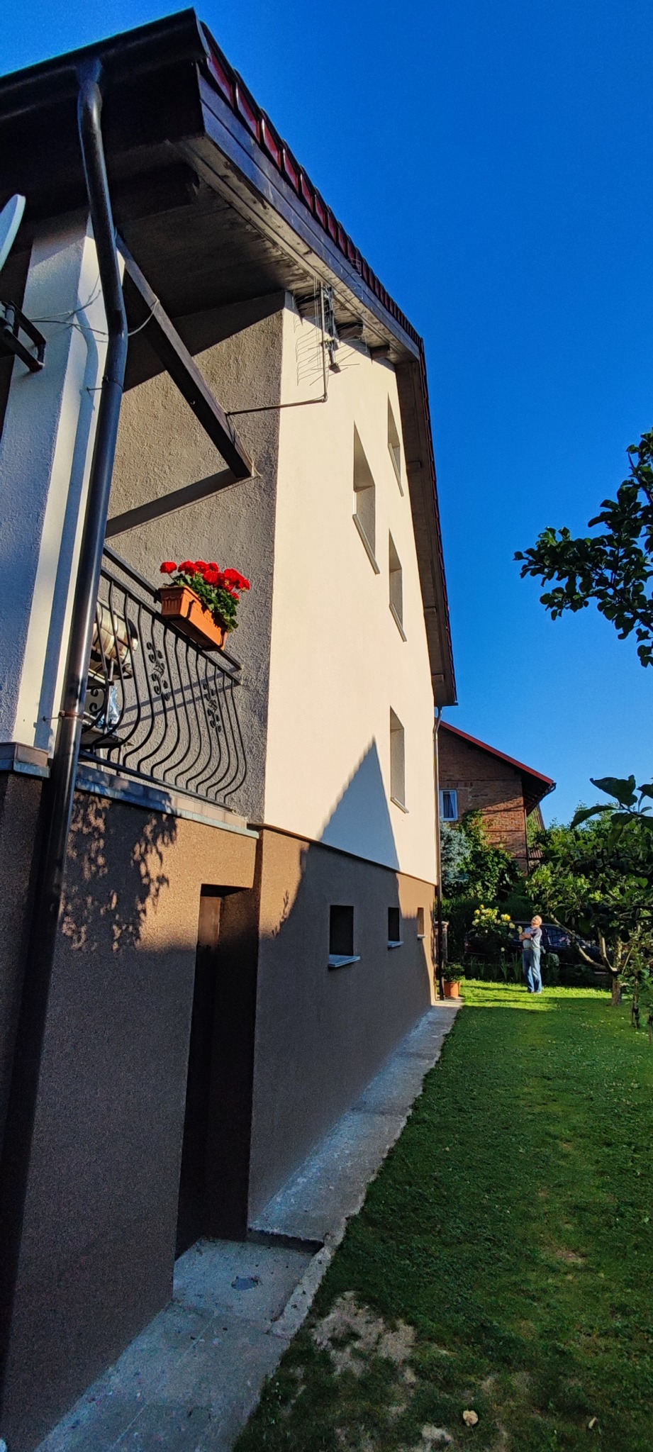 Dwukondygnacyjny dom z jasną elewacją i ciemnobrązowym cokołem, balkon z czerwonymi kwiatami, widok z dołu na tle błękitnego nieba i zielonego trawnika.