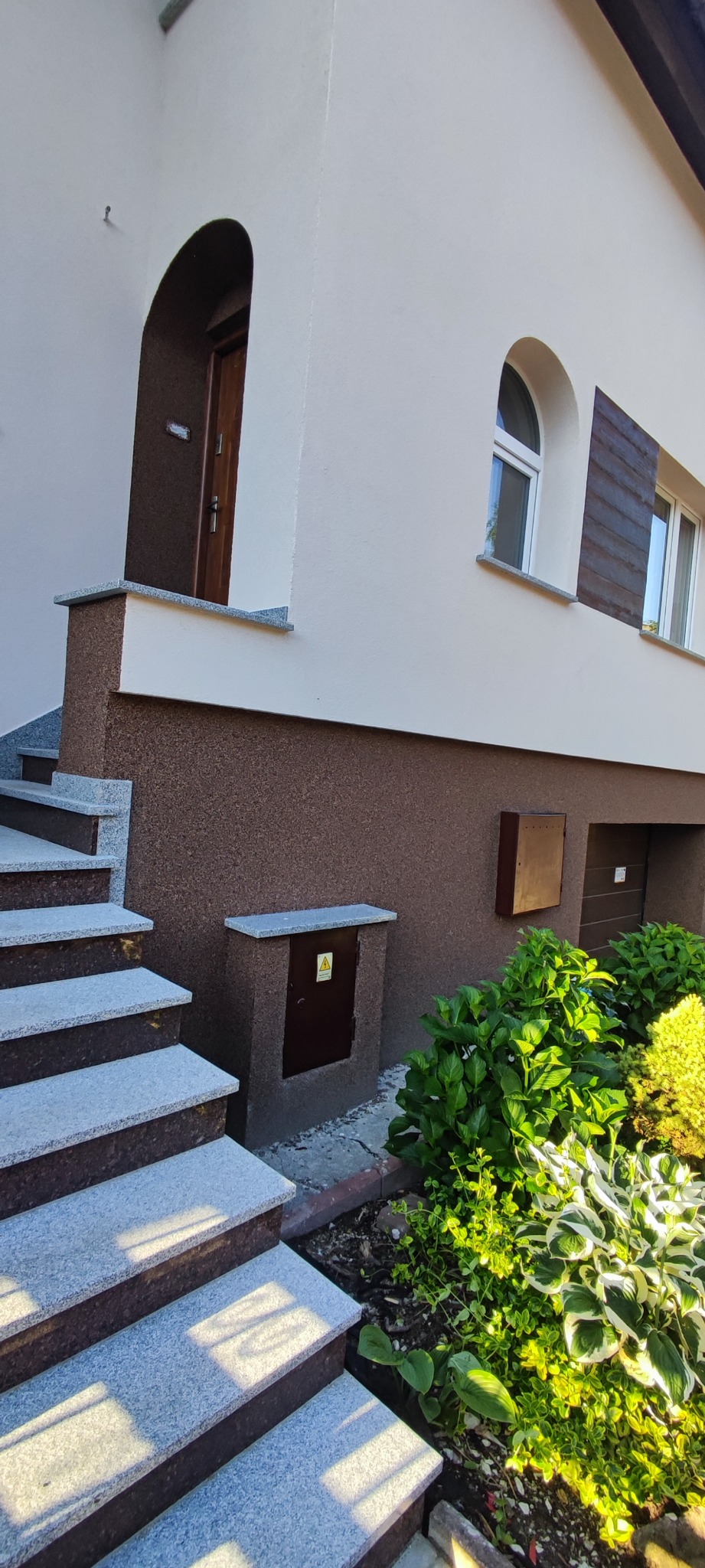 Dwukondygnacyjny dom z elewacją w odcieniach beżu i brązu, z granitowymi schodami wejściowymi i małą skrzynką elektryczną z symbolem ostrzegawczym.