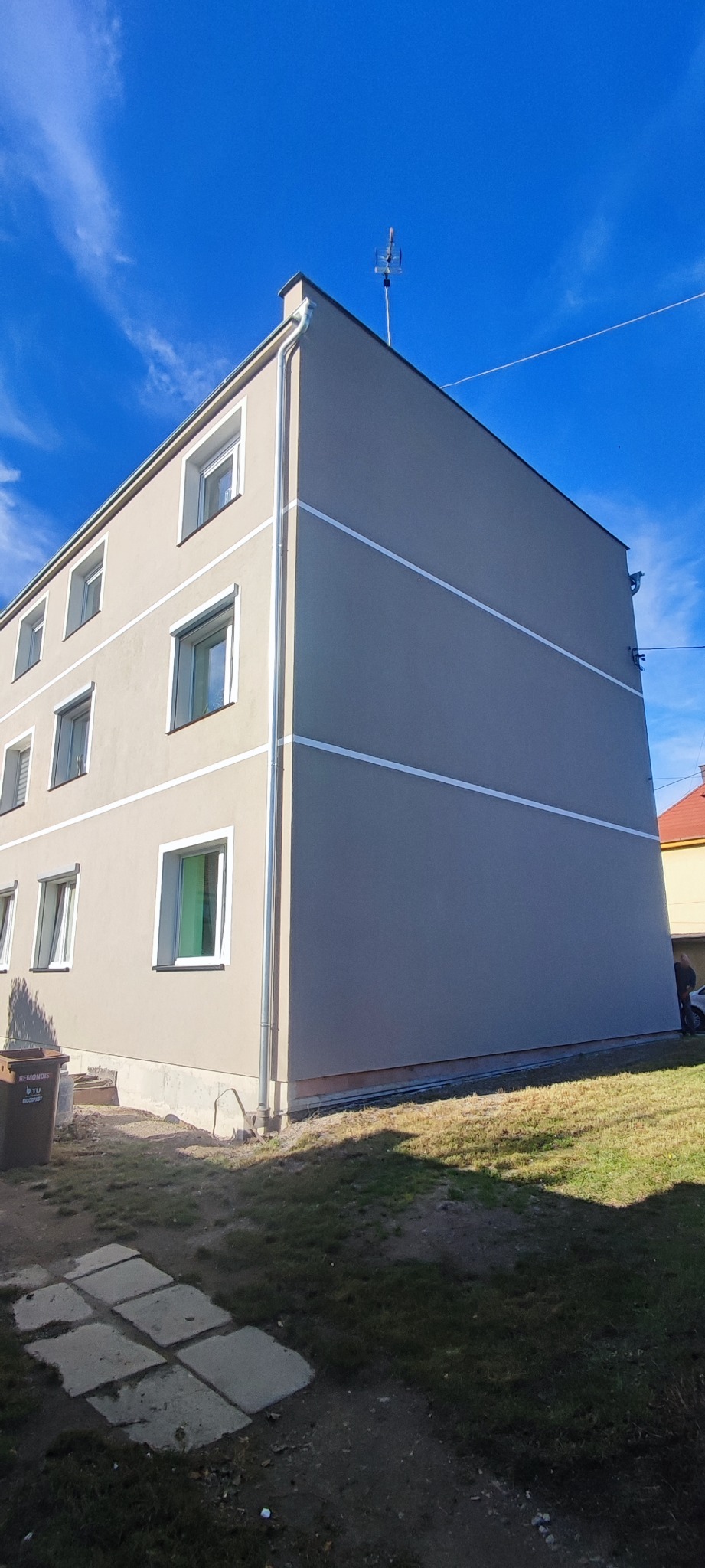 Odnowiony budynek wielorodzinny z szarą elewacją, białymi pasami i nowymi oknami, widoczny z poziomu gruntu na tle błękitnego nieba.