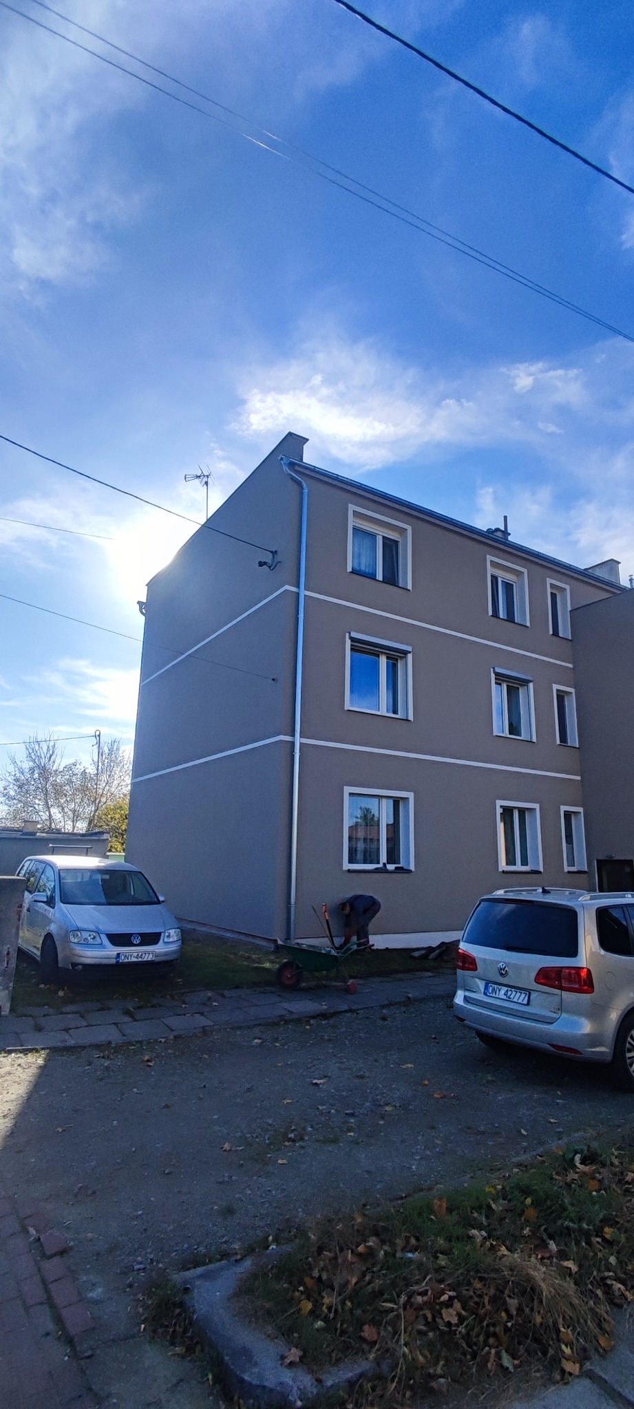 Odnowiony budynek wielorodzinny z szarą elewacją i białymi pasami, widoczne nowe rynny, pracownik z taczką i narzędziami przy budynku, dwa samochody na podwórku, błękitne niebo.