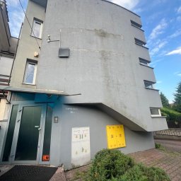 ALIGATOR Alicja Gatlik - Szara, wielopiętrowa elewacja budynku z charakterystycznym zaokrągleniem i widocznymi śladami zabrudzeń, z wejściem z ciemnymi drzwiami, skrzynkami gazowymi i elektrycznymi oraz roślinnością u...