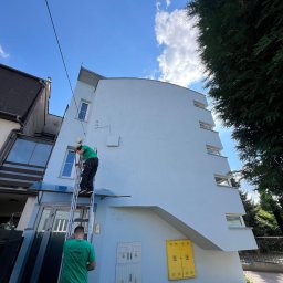 ALIGATOR Alicja Gatlik - Pracownik na drabinie, w trakcie odświeżania koloru elewacji budynku w Myślenicach, widoczne puszki z farbą i narzędzia malarskie.