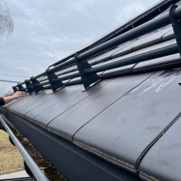 ALIGATOR Alicja Gatlik - Czyszczenie rynny dachowej z nagromadzonych liści, widoczne metalowe zabezpieczenie przed śniegiem, dachówka ceramiczna i fragment dłoni pracownika.