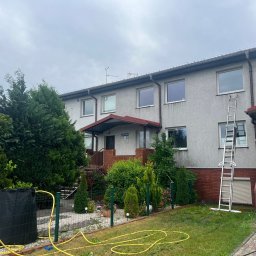 ALIGATOR Alicja Gatlik - Dwupiętrowy budynek z szarą elewacją, widoczną drabina oparta o ścianę, żółty wąż ogrodowy leżący na trawniku, oraz zbiornik na deszczówkę w ogrodzie.