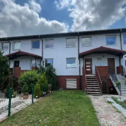 Szereg domów z szarą elewacją, brązowymi rynnami i czerwonymi ceglanymi elementami, widok z poziomu trawnika w pochmurny dzień.