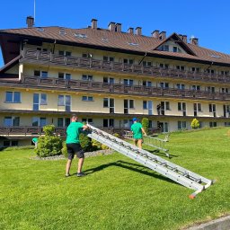 ALIGATOR Alicja Gatlik - Dwóch mężczyzn w zielonych koszulkach transportuje rozłożoną drabinę na trawniku przed trzypiętrowym budynkiem z brązowym dachem i balkonami.