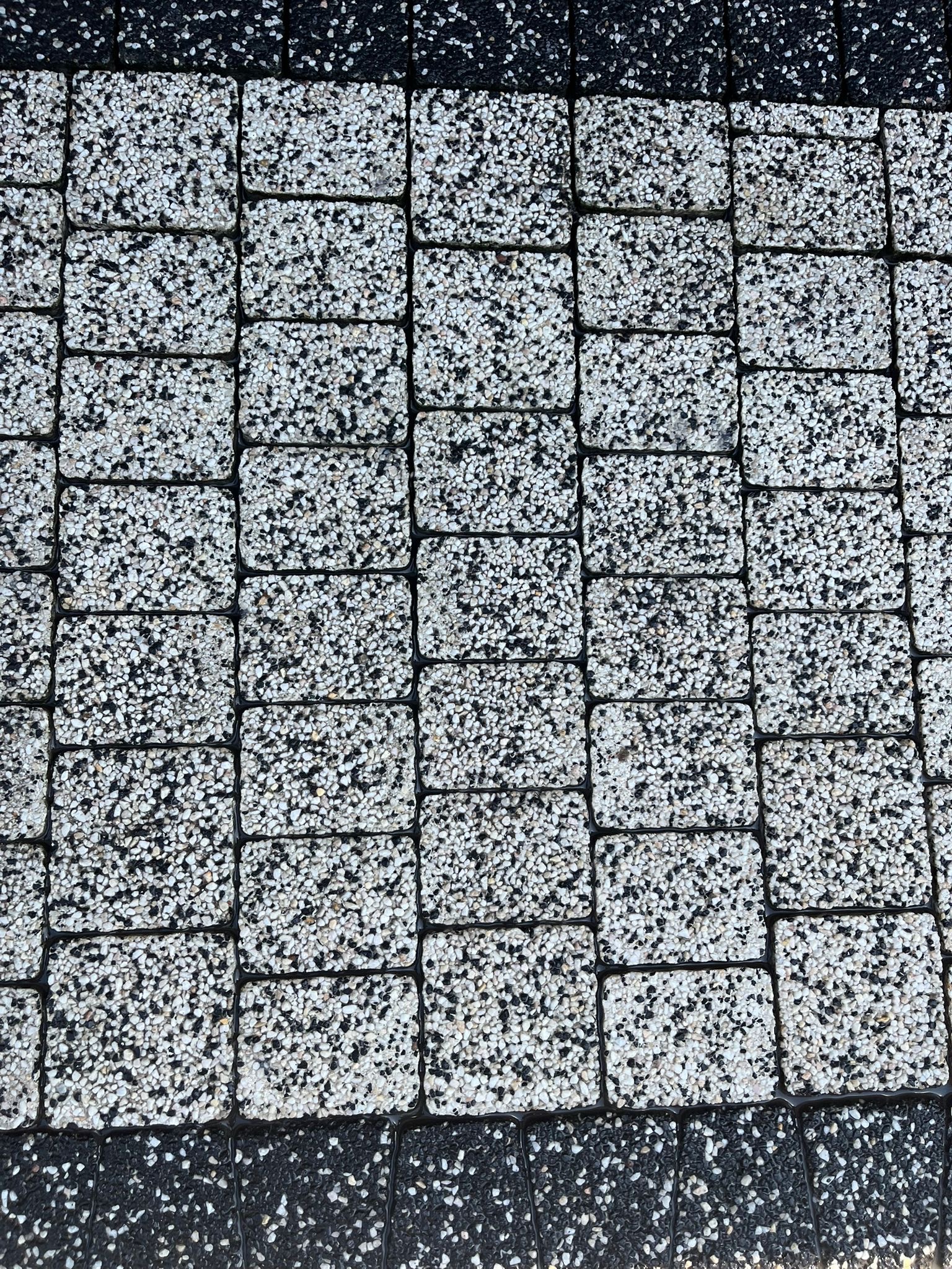 Detale ułożonej kostki brukowej z drobnymi kamyczkami w odcieniach bieli i szarości, obramowanej czarną kostką, tworzącej kontrastowy wzór.