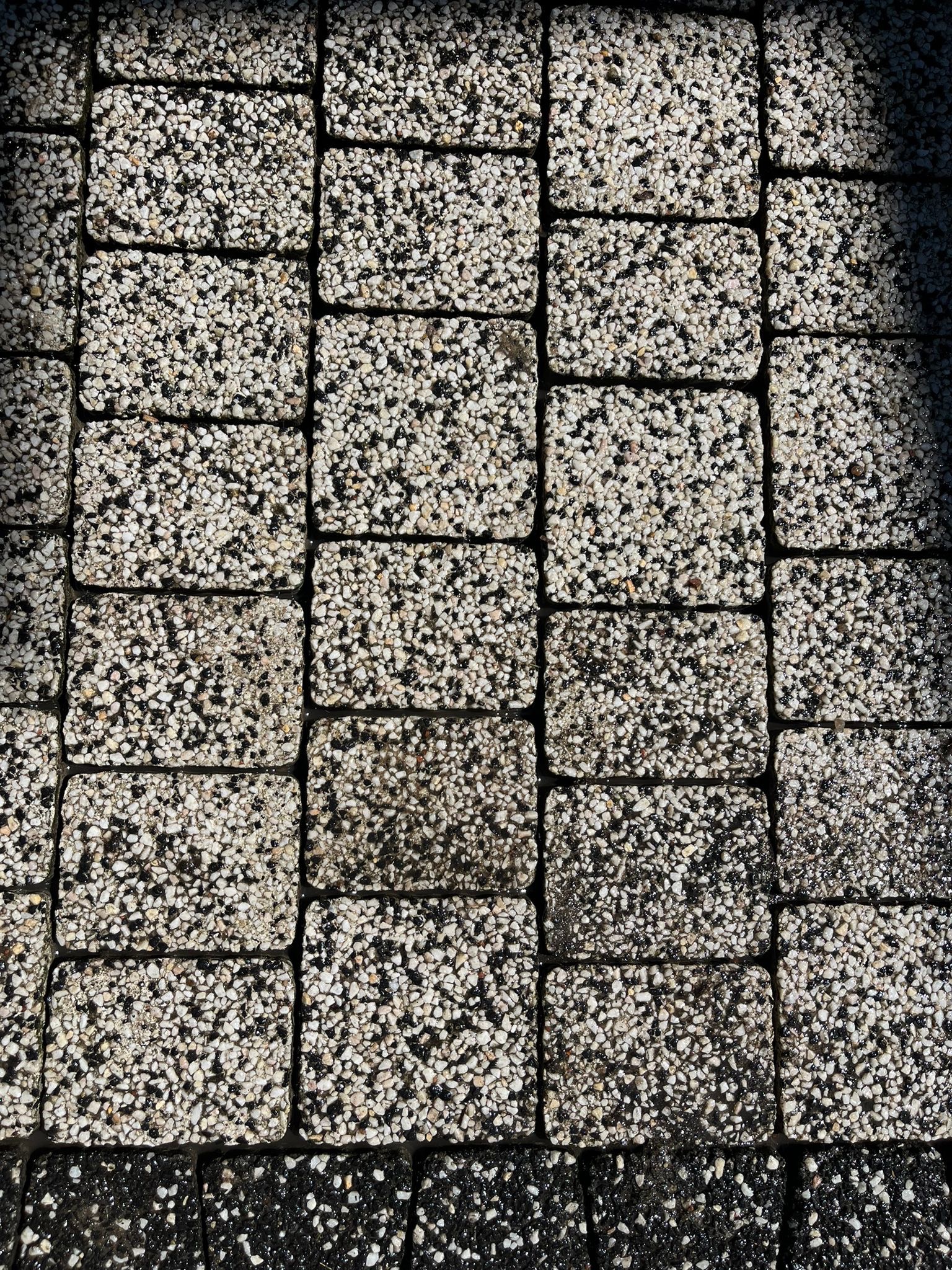 Zbliżenie na ułożoną kostkę brukową z drobnymi kamyczkami, widoczne różnice w odcieniach i fakturze poszczególnych elementów, z delikatnym cieniem po prawej stronie.