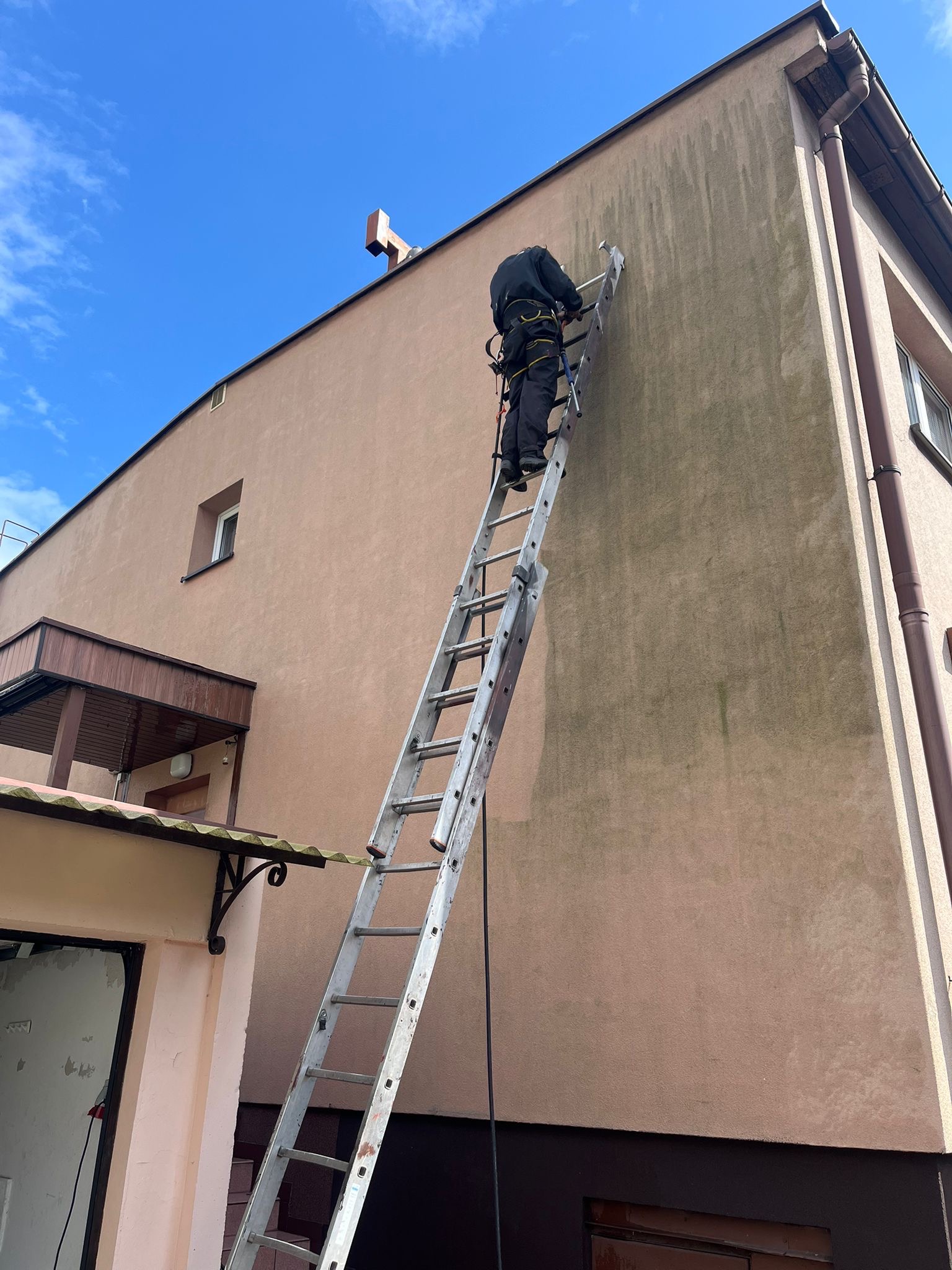 Pracownik w uprzęży na drabinie podczas mycia zabrudzonej elewacji budynku z beżowym tynkiem.