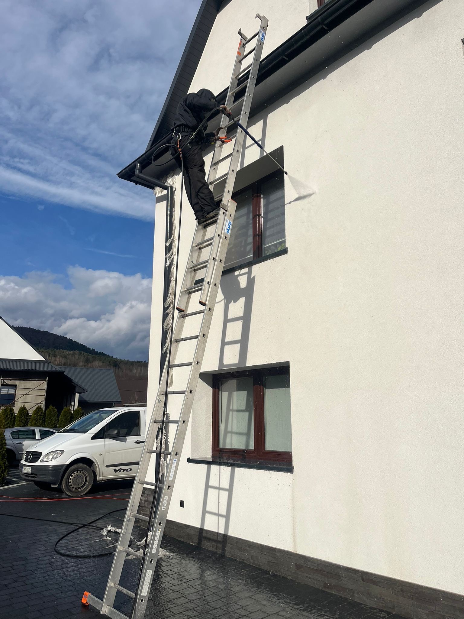 Pracownik na drabinie czyści elewację budynku pod ciśnieniem, widoczny spływający brud, w tle biały samochód dostawczy i błękitne niebo.