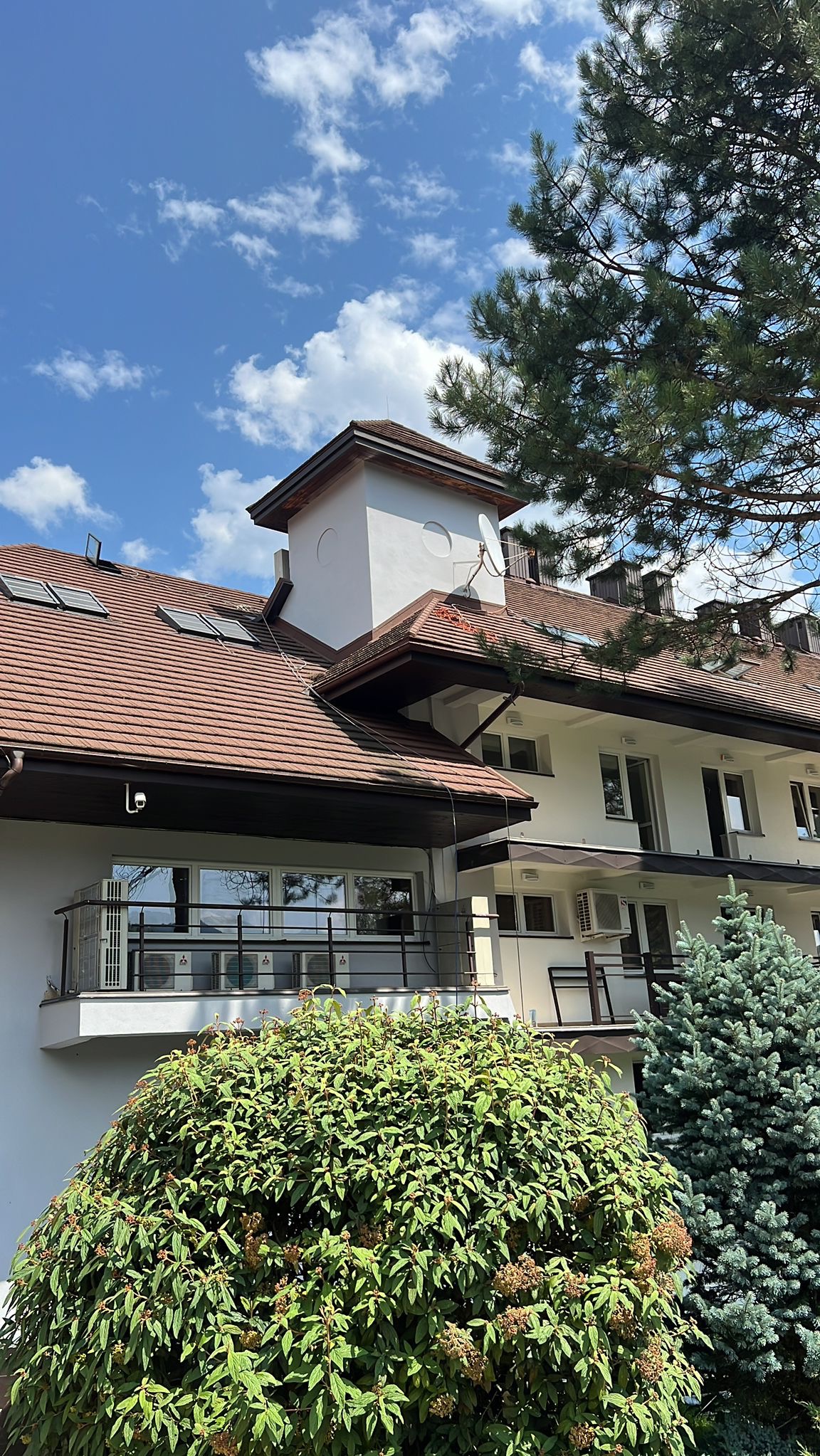Wielopiętrowy budynek z balkonem, brązowym dachem z lukarnami i białymi ścianami, widoczny z poziomu gruntu, z bujną zielenią w pierwszym planie i błękitnym niebem w tle.