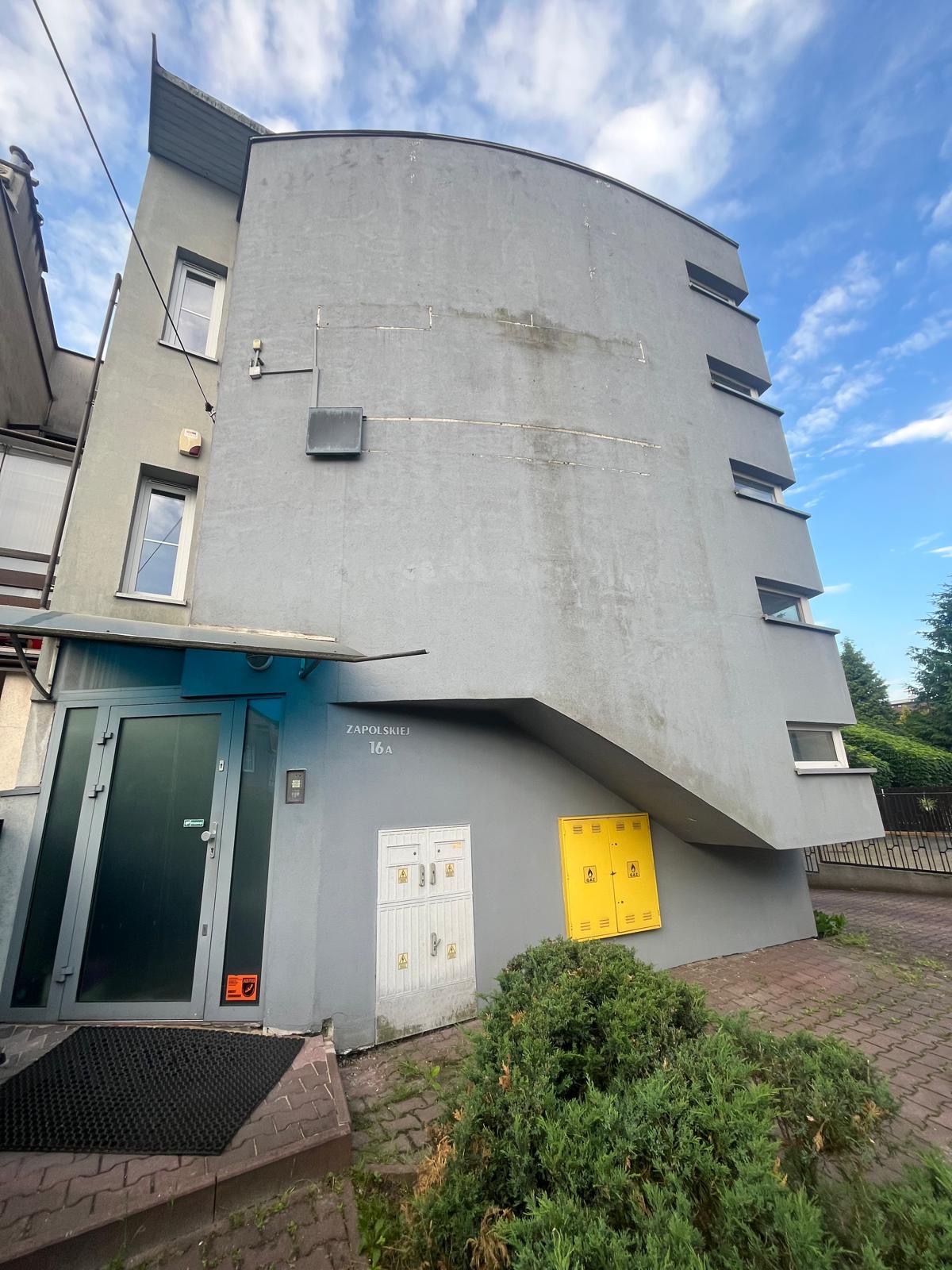 Szara, wielopiętrowa elewacja budynku z charakterystycznym zaokrągleniem i widocznymi śladami zabrudzeń, z wejściem z ciemnymi drzwiami, skrzynkami gazowymi i elektrycznymi oraz roślinnością u...