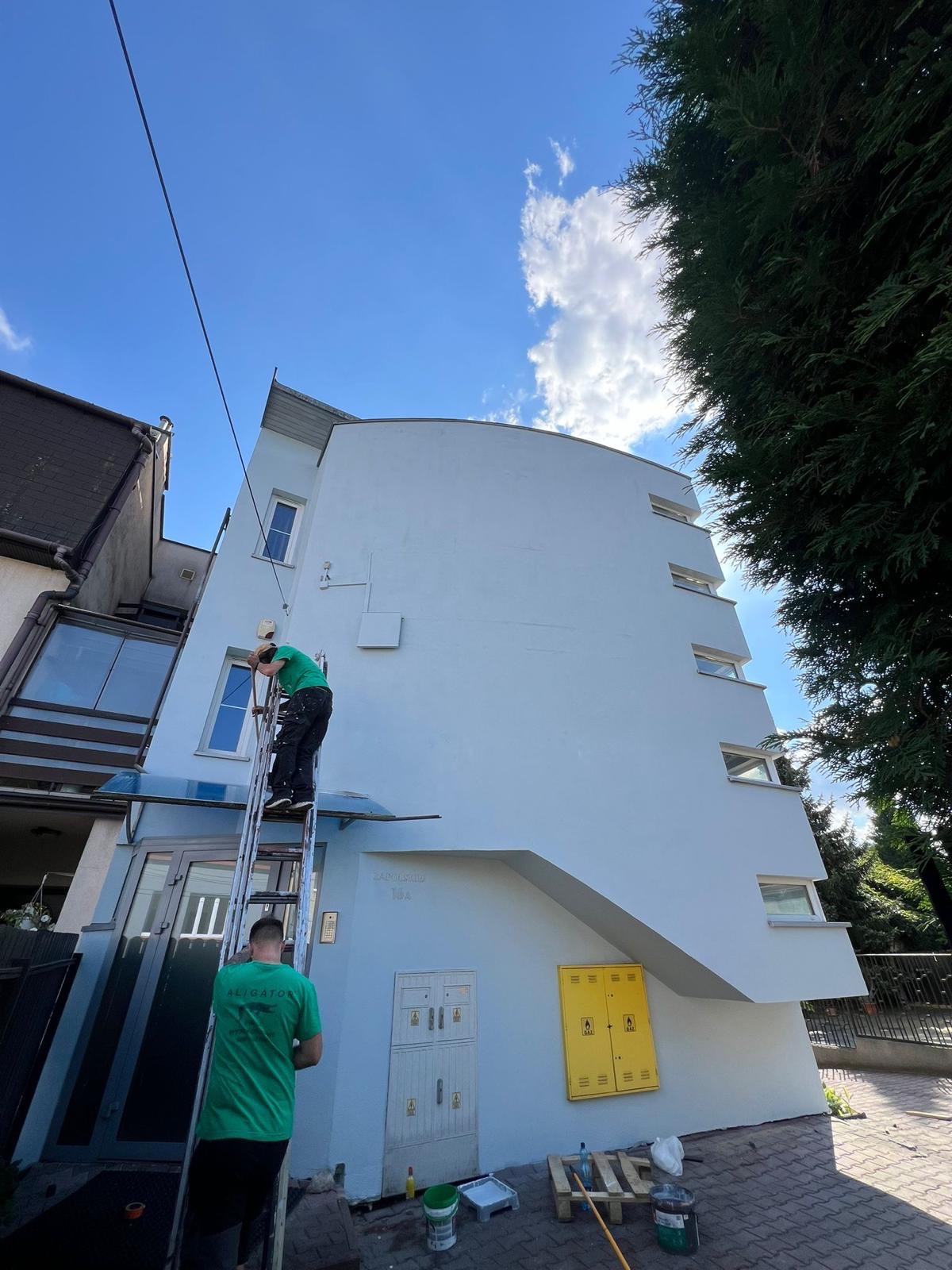 Pracownik na drabinie, w trakcie odświeżania koloru elewacji budynku w Myślenicach, widoczne puszki z farbą i narzędzia malarskie.